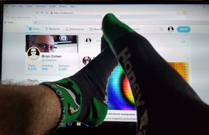 Feet wearing green and black Hohey socks rest in front of a laptop displaying a Twitter page for Brian Cohen (@nthepixoils).
