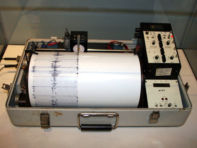 A seismograph machine in a metal carrying case displaying a recorded seismic wave pattern on a paper drum printout.