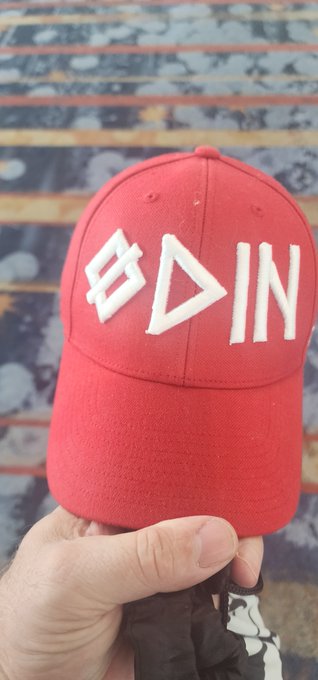 A hand holds up a red baseball cap with white embroidered symbols resembling the Bitcoin logo and geometric shapes, positioned in front of an American flag background.