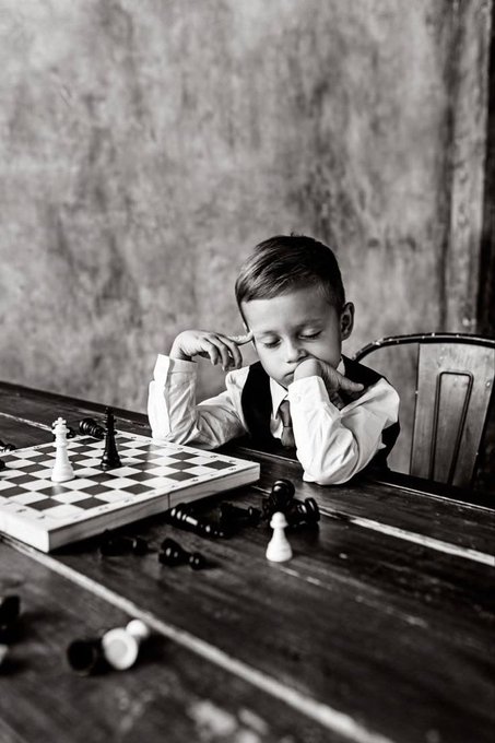 A black and white photograph of a young boy in a vest and tie sitting at a wooden table, thoughtfully resting his chin on his hand while looking at a chessboard with scattered pieces.
