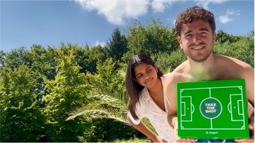 Two people outdoors near greenery, with the person on the right holding a green card labeled 'MAKE YOUR LUCK'.
