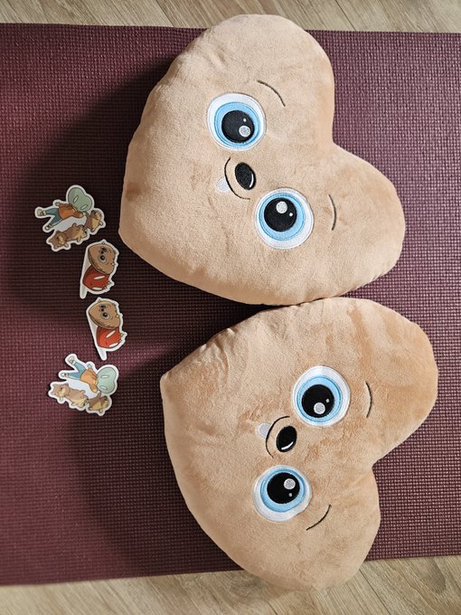 Two peanut-shaped plush cushions with cartoon faces and big blue eyes, alongside small paper cutouts of insects and a peanut character on a maroon mat.