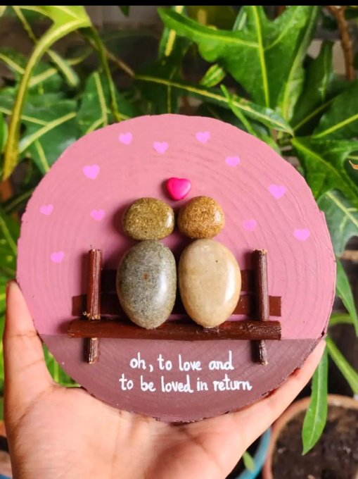 A hand holds a round pink decorative plaque featuring two pebbles arranged to look like a couple on a bench, with the text 'oh, to love and to be loved in return' written below.