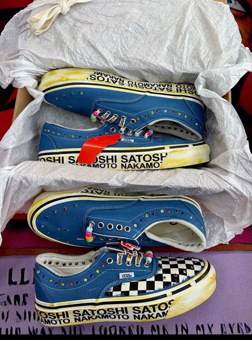 Two pairs of blue Vans sneakers featuring custom artwork of Bitcoin creator Satoshi Nakamoto are displayed in their tissue-lined boxes, with "SATOSHI NAKAMOTO" printed along the white soles and "VANS" logos on the sides. The collaborative footwear, adorned with colorful rhinestones and checkerboard patterns on the toe caps, represents the exclusive release causing excitement and "fights" at ComplexCon.