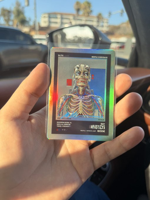 A hand holds up a holographic Beeple "Everydays" NFT trading card (serial #18101) with an error print visible on the front, taken from inside a car with palm trees and buildings in the background, perfectly illustrating the "Beeple error card coming home" caption.
