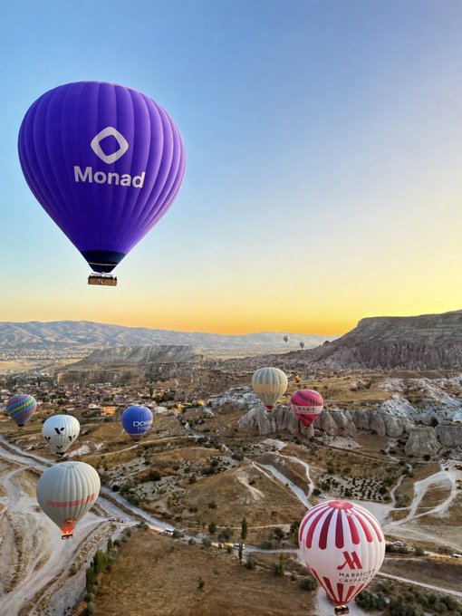 A purple hot air balloon with the 'Monad' logo and name floats above a landscape at sunset, with several other balloons visible in the distance over a rocky terrain resembling Cappadocia.