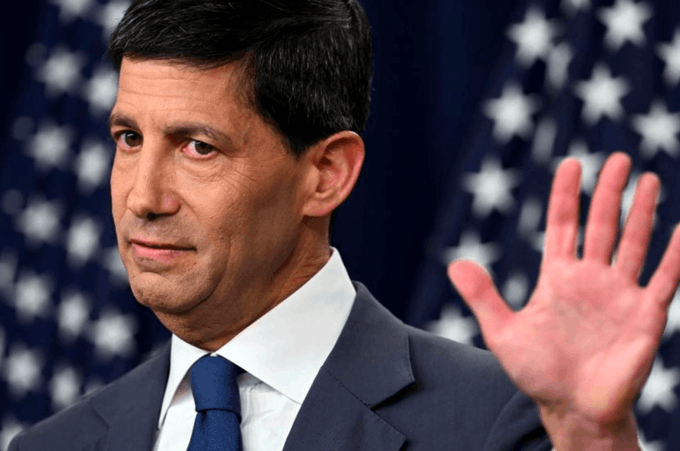 A man in a dark blue suit and tie raises his left hand with a serious expression, standing in front of two blurred American flags.