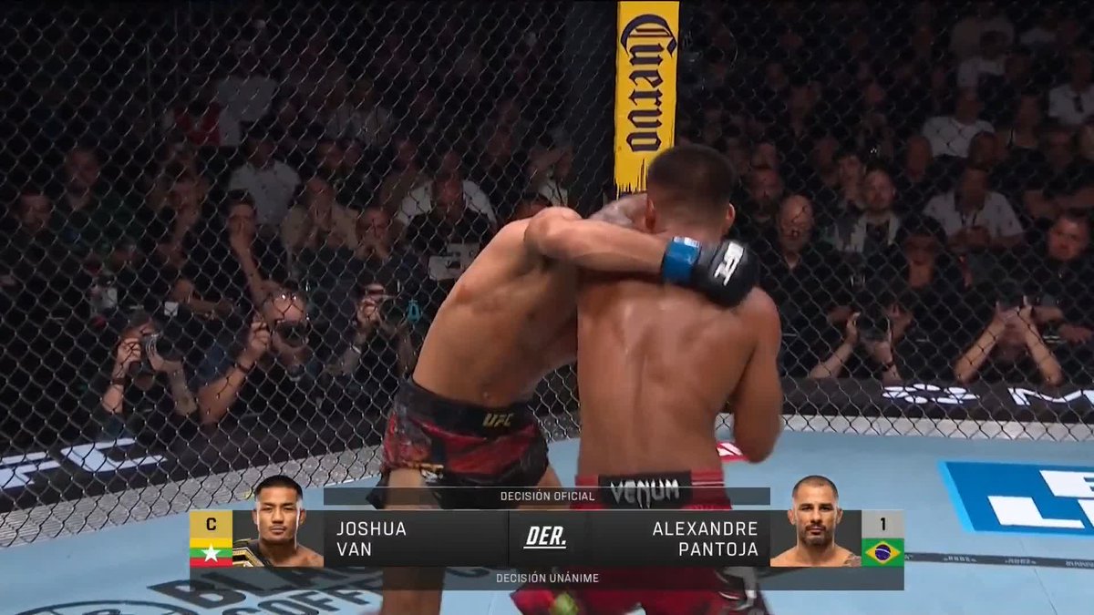 Two UFC fighters embrace in the octagon after a championship bout, with an on-screen graphic declaring "Joshua Van" the winner by "DECISION UNÁNIME" over "Alexandre Pantoja." The image captures the post-fight moment following Van's successful title defense, as referenced in the accompanying social media post.