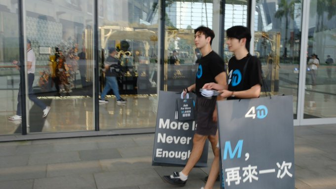 Two young men in black T-shirts stand beside large promotional signs at the entrance of a modern glass building, apparently promoting 4MEXchange. Visible text includes “4D” on the shirts and sign, “More Is Never Enough,” and partial Chinese text including “M, 再来一次,” matching the post’s point about using eye-catching male models and bold signage to draw attention at a busy exhibition.
