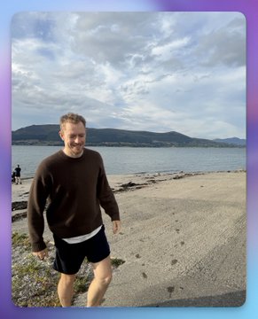 A man in a brown sweater and black shorts jogging along a beach with mountains and water visible in the background.