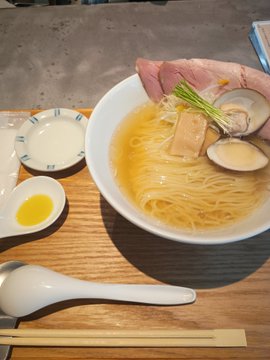A bowl of shio (salt) ramen with clams (asari), pork slices, and green onions, served with a soft-boiled egg on the side at a restaurant.
