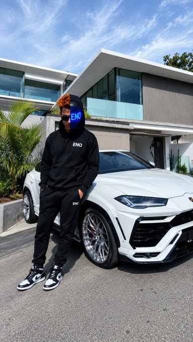 A person wearing a black "ENO" hoodie and matching track pants stands next to a white Lamborghini SUV in front of a modern house. They are wearing a distinctive blue mask with the "ENO" logo and sunglasses.