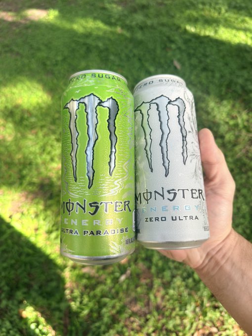 A hand holds two Monster Energy drink cans, a green "Ultra Paradise" and a white "Zero Ultra", in front of a grassy background.