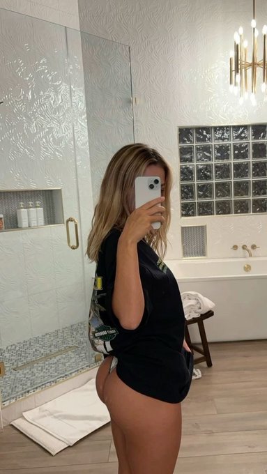 A person with long blonde hair takes a mirror selfie in a modern bathroom featuring a glass-enclosed shower, a bathtub with a glass block window, and a contemporary chandelier.