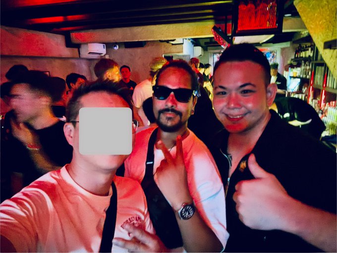 A photo of three men posing together in what appears to be a bar or club setting; one face is obscured with a gray rectangle, the middle man wears sunglasses, and the rightmost man gives a thumbs-up.