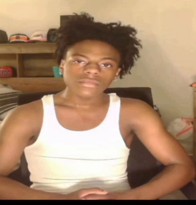 A young Black man with curly hair wearing a white tank top, sitting indoors with a wooden shelf displaying caps behind him.