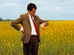 A man in a brown jacket, white shirt, and red tie stands in a field of yellow flowers, checking his wristwatch.