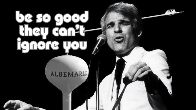 A black-and-white photo of a man in a white shirt and tie speaking into a microphone, with the text "be so good they can't ignore you" and a white water tower labeled "ALBEMARLE".