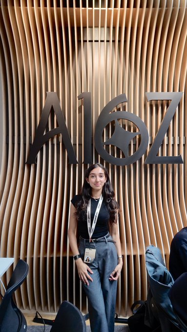 A young woman with long dark hair poses confidently in front of a large "A16Z" logo displayed on a wooden slatted wall, wearing a lanyard badge that suggests her participation in the a16z accelerator program referenced in the post. The image captures the professional, in-person moment at a16z's offices that complements her advice about navigating the accelerator experience.