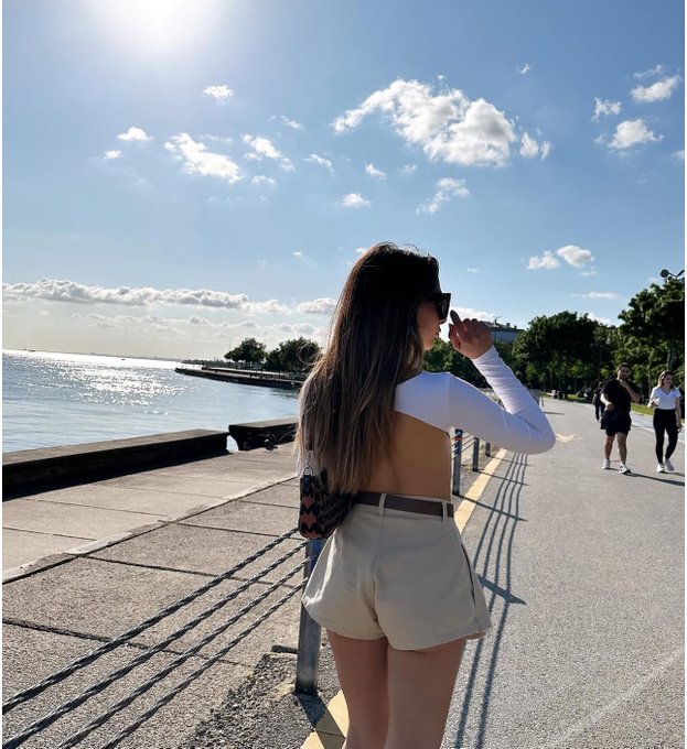 The image shows a woman walking down a sidewalk near water, possibly at the beach or by the ocean. She has her hand up to her face as if she's taking a photo of something interesting or important. The surrounding area appears to be a popular spot for people to enjoy the view and take pictures.