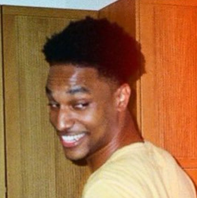 A young man with a high-top fade haircut smiles and looks back over his shoulder in a warmly lit room with wooden doors.