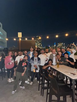 Group photo of people gathered at an outdoor rooftop bar at night with city skyline and string lights in the background.