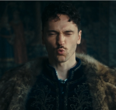 A close-up of a man with dark hair and a beard wearing an ornate dark tunic with fur trim, appearing to be a scene from a historical or fantasy film or TV series.