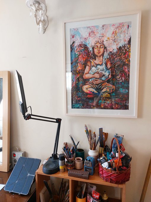 An artist's workspace featuring a desk lamp, paintbrushes, and various art supplies, with a colorful framed painting of a seated figure hanging on the wall above.