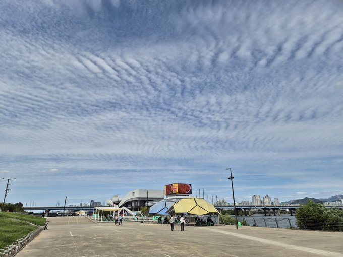 An outdoor scene under a sky filled with rippled altocumulus clouds, showing a waterfront promenade, colorful tent-like structures, and a city skyline across the water.