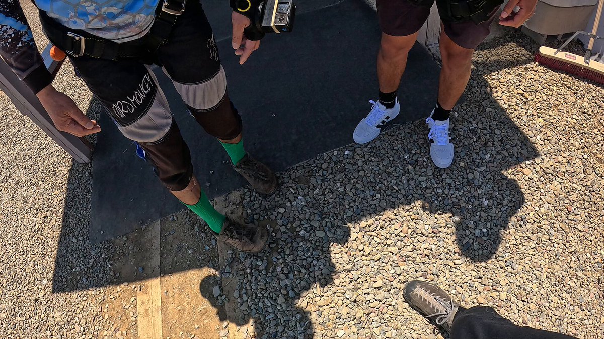 The image features two people standing next to each other, both wearing black shorts with green socks. One person has their feet propped up on a wall or ledge while the other person's feet are resting on the ground. The background of the photo is blurred, but there appears to be gravel and dirt present in the scene.

The image also contains text that reads "Bro literally took