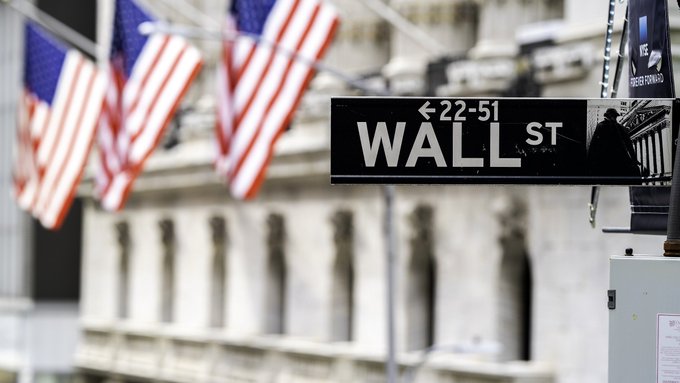 A Wall Street sign marked “WALL ST” and “22–51” stands in front of blurred American flags and a building facade. It visually anchors the post’s discussion of Binance bStocks expanding from five U.S. stocks in June to 100 listings by 21 October, including major companies and indexes.