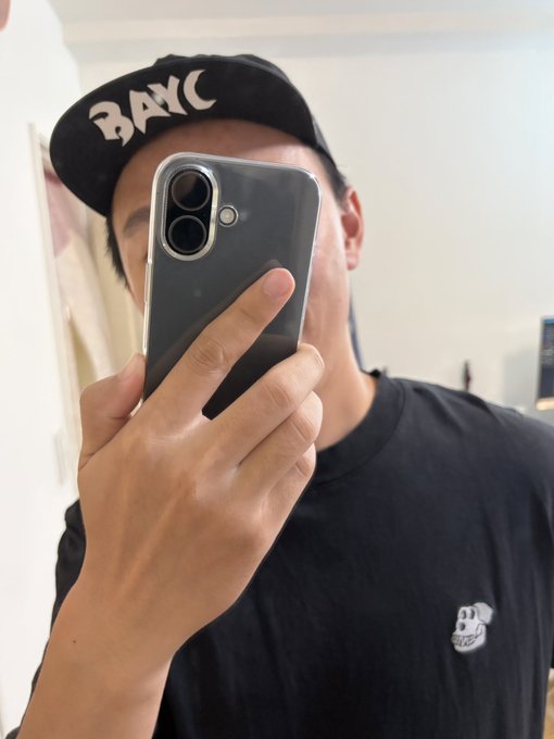 The image features a man taking a selfie with his phone, wearing a black shirt that has a small dog design on it. The background of the photo shows a white wall, which contrasts nicely with the black shirt and the man's face. The caption "@BoredApeYC" is also present in the image, possibly indicating the account or the person who posted this selfie.