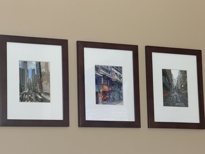 Three dark wooden picture frames hang on a beige wall, each containing a different urban cityscape photograph with wide white mats.