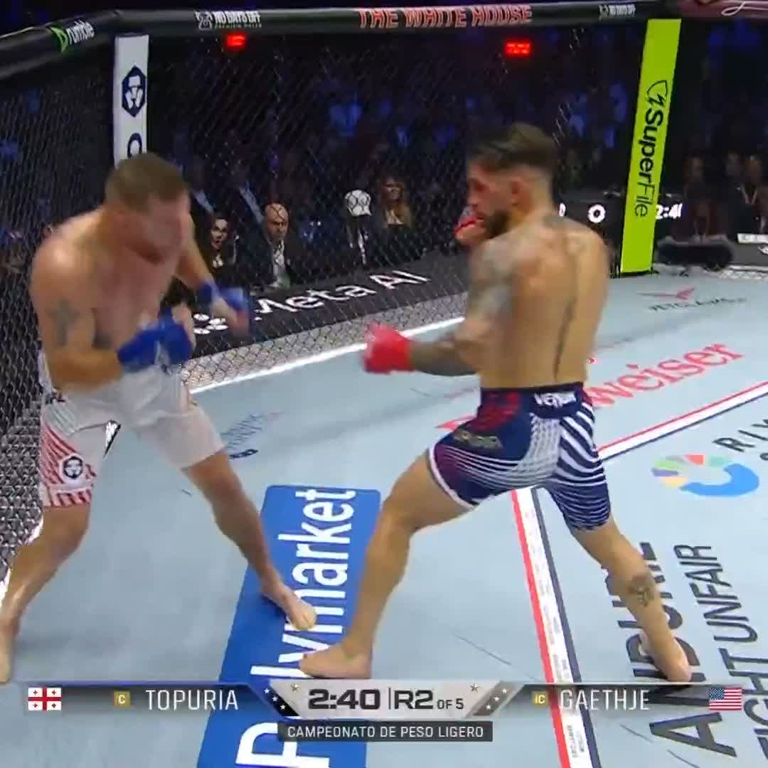 The image shows two boxers fighting each other inside a ring, with one boxer wearing white shorts and the other wearing blue shorts. The fighter in white shorts has his mouth open, while the fighter in blue shorts appears to be throwing a punch at him. Both boxers are wearing gloves as they engage in this intense match. In the background, there is a crowd of spectators watching the fight intently from behind a fence.