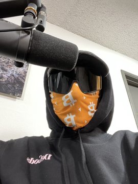 A person wearing a black hoodie, sunglasses, and an orange bandana covering their face stands in front of a microphone.