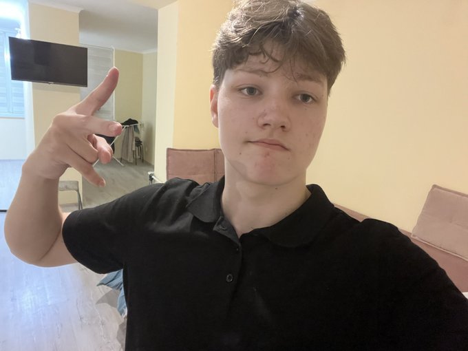 A teenage boy in a black polo shirt takes a selfie indoors, pointing his finger upward with a TV and living room visible behind him.