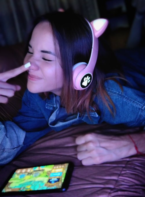 A young woman lies on a bed with her eyes closed, wearing pink cat-ear headphones while a smartphone displays a colorful mobile game in the foreground, evoking a moment of immersive gaming that ties into the post's nostalgic celebration of Axie Classic. The relaxed, content expression mirrors the heartfelt affection the text expresses toward the game that sparked her journey into the gaming world.