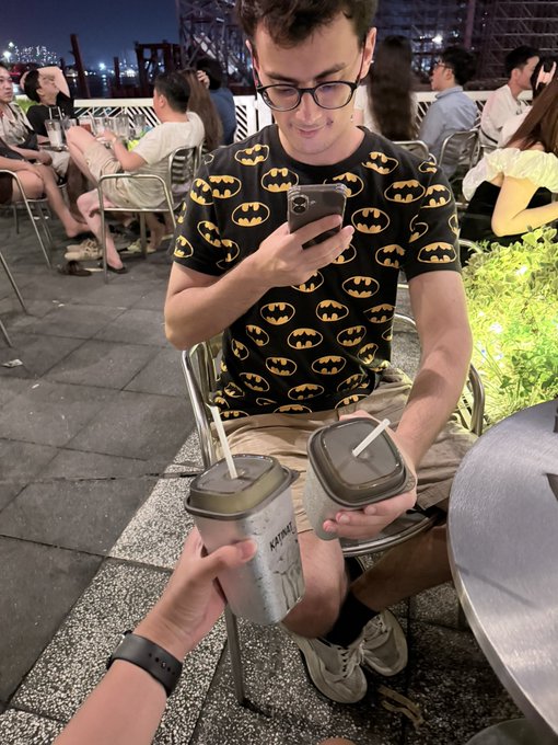 A young man wearing glasses and a Batman-logo patterned shirt sits at an outdoor café looking at his phone while holding two drinks with straws, as a first-person hand extends another drink toward him. One cup reads "KAPIME" and the post jokingly frames this coffee meetup as a "date with the most special person in the @monad community" where they're celebrating hopes that $MON will reach $1.