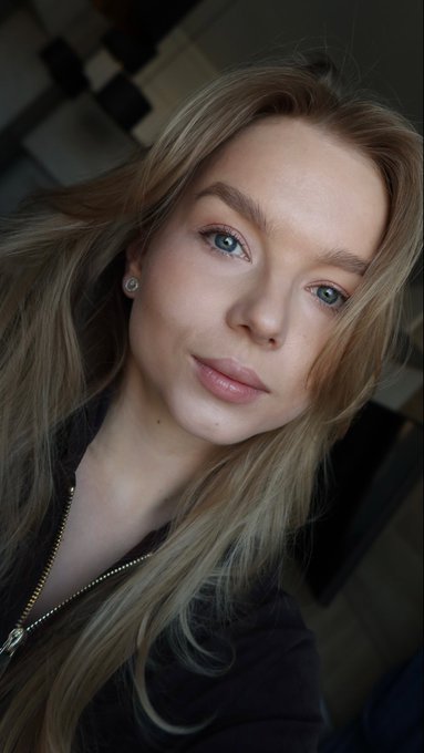 A close-up selfie of a young woman with long blonde hair, blue eyes, and pink lipstick, wearing a small stud earring and a dark zip-up jacket.
