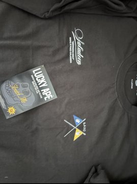 A black T-shirt is displayed with a luxury ape BAYC x King Saladeen tag still attached, featuring "Quality" text and "Greatest of All Time" embroidery near the collar, along with a small colorful logo. The image matches the post's description of a newly received Bored Ape Yacht Club collaboration T-shirt with attached branding tag.