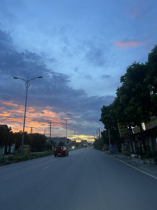 This is a nighttime photo of an empty street with some cars driving down it, including one car that appears to be parked by the side of the road. The sky has an orange hue at sunset, giving the scene a warm ambiance as the sun sets over the landscape.