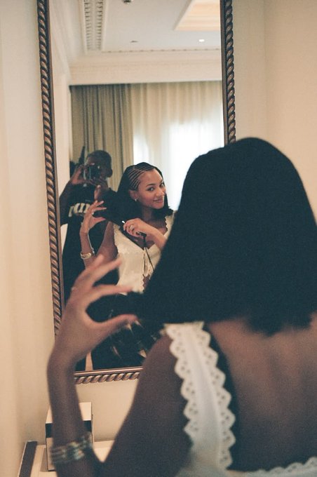 A woman takes a mirror selfie in an elegant hotel room while another person in the background photographs her with a camera.