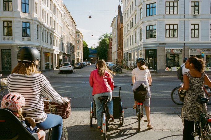 Several people, including women with helmets and a child in a pink helmet, ride bicycles down a cobblestone street lined with European-style buildings on a sunny day.