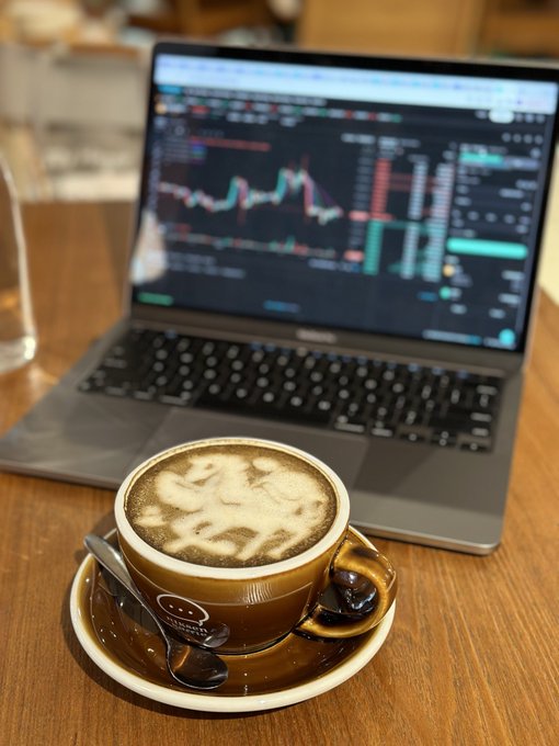 A cappuccino with latte art sits on a wooden table in front of an open laptop displaying a financial trading chart with candlestick patterns.