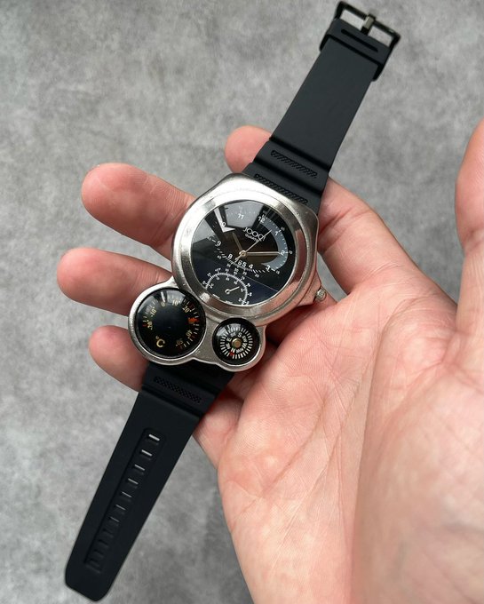 A hand holds a unique dual-dial watch with a silver case, black face, and an additional small sub-dial featuring colorful zodiac symbols, attached to a black rubber strap.
