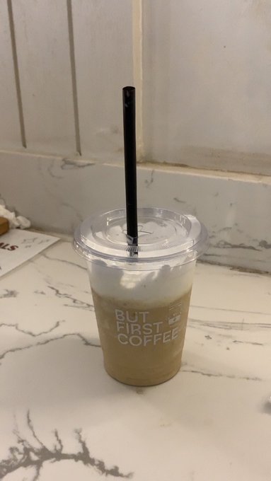 An iced coffee in a clear plastic cup with a black straw sitting on a white marble surface, with 'BUT FIRST COFFEE' printed on the cup.