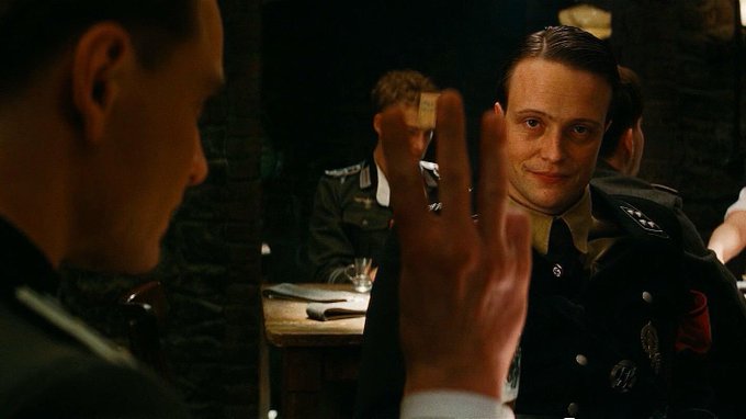 Scene from the film 'Inglourious Basterds' showing a German officer holding up three fingers, with another officer visible in the background mirror.