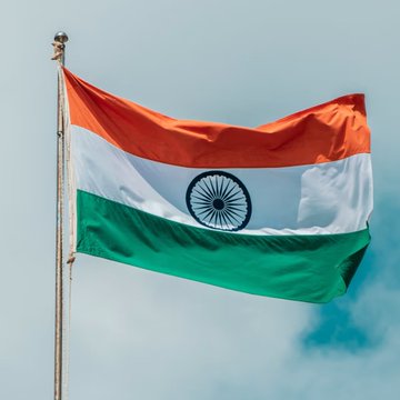 The image shows the Indian national flag, featuring its saffron, white, and green horizontal stripes with the navy blue Ashoka Chakra in the center, waving against a cloudy sky. The flag directly relates to the post by representing India, the country whose financial market the Binance APAC Head is commenting on regarding the need for rupee stablecoins to mitigate US dollar volatility.