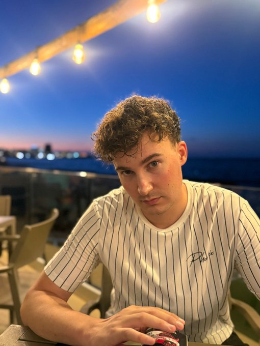 A young man with curly brown hair wearing a white pinstriped shirt looks at the camera with a serious expression, seated at an outdoor table with string lights and a sunset visible in the background.