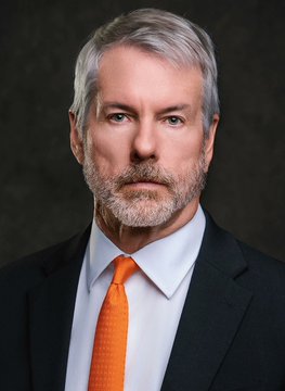 A distinguished man with gray hair and a beard, wearing a dark suit, light blue shirt, and an orange tie, gazes directly at the camera. The orange tie likely symbolizes his bullish stance on Bitcoin, aligning with the post's reference to him "waving the orange flag" and signaling an imminent BTC purchase for Strategy (MSTR), which already holds 845K+ BTC worth $68.5B.